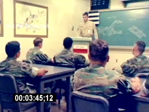 The still from the 'USMC Aviation Ordnance Recruiting Film - Silent' depicts a classroom scene featuring a military instructor standing at a podium, addressing a group of seated recruits. The recruits, dressed in camouflaged uniforms, focus intently on the presentation, which likely involves educational materials displayed on a chalkboard. Maps and diagrams related to aviation ordnance are visible on the walls, emphasizing the instructional nature of the environment. The overall atmosphere appears serious and focused, highlighting the recruitment and training process in the Marine Corps.
