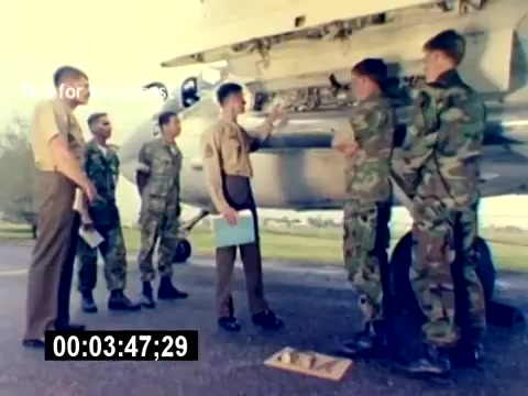 The still depicts a training scene from the USMC Aviation Ordnance Recruiting Film. It features a group of military personnel, including a uniformed instructor and recruits in camouflage, gathered around an aircraft. The instructor appears to be explaining or demonstrating something related to aviation ordnance. The setting is outdoors, likely on an airfield, emphasizing the hands-on training aspect of the program.