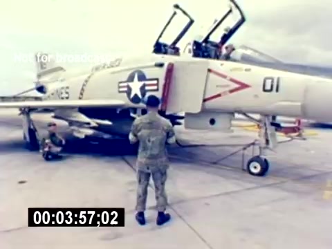 The still features a military aircraft, likely a jet, with visible markings including the U.S. Navy star insignia and identification numbers. Two service members are engaged with the aircraft; one stands near the front, possibly preparing for maintenance or inspection, while another is positioned underneath. The setting appears to be a military airfield, emphasizing the operational aspects of U.S. Marine Corps aviation. The scene lacks audio, indicating a focus on visual elements that showcase aviation ordnance and recruitment.
