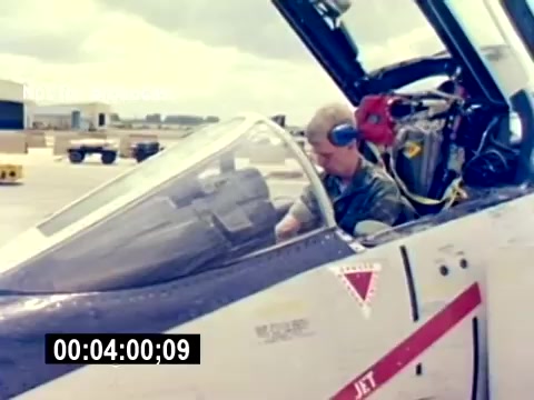 The still shows a U.S. Marine Corps aircraft, specifically a fighter jet, with a service member inside the cockpit. The individual appears focused, possibly preparing for takeoff or conducting pre-flight checks. Surrounding the jet are features of an airbase, with a clear sky and open space visible in the background. The overall atmosphere conveys a sense of duty and the technical nature of military aviation.