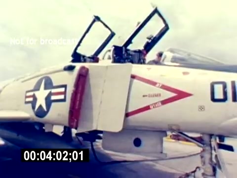The still shows a military aircraft, likely a fighter jet, with its cockpit canopy open. The plane is adorned with a star insignia and red accents, indicative of U.S. Marine Corps colors. The focus is on the cockpit area, where personnel appear to be preparing for operations. The timestamp suggests this is part of a longer film. The overall context hints at the film’s purpose of recruiting and showcasing aviation ordnance roles within the USMC.