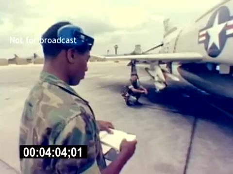 The still from the 'USMC Aviation Ordnance Recruiting Film - Silent' depicts two individuals on an airfield, focusing on a military aircraft. One person, wearing a camouflage uniform and a cap, is holding a clipboard, likely reviewing maintenance or ordnance details. The other individual appears to be inspecting or working on the aircraft, which features a distinct military insignia. The environment suggests a hangar or tarmac setting, typical for aviation operations. The overall tone is professional, emphasizing the technical and operational aspects of aviation ordnance work.
