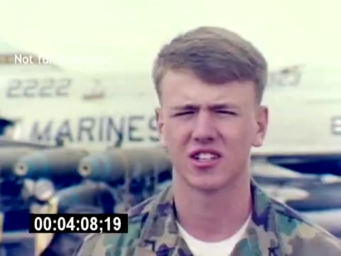 The still features a young male Marine in camouflage uniform, positioned in front of military aircraft, likely featuring ordnance. He appears to be speaking directly to the camera, conveying information related to his role or experiences. The background shows military planes, emphasizing a recruiting context focused on USMC Aviation Ordnance. The timecode suggests it is part of a longer film sequence.