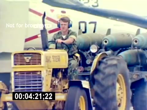 The still shows a service member in military attire driving a yellow tractor on an airfield, with aircraft visible in the background. The tractor is towing ordnance, likely bombs or ammunition. The scene emphasizes the logistics and support role in aviation ordnance operations within the US Marine Corps. The absence of sound suggests a focus on visual elements, highlighting the working environment.
