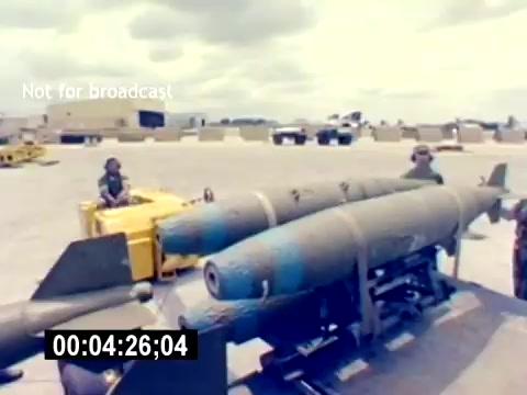 The still from the 'USMC Aviation Ordnance Recruiting Film - Silent' features two military personnel near a set of bombs on a transport vehicle. The bombs are prominently displayed, showcasing their cylindrical shape and markings. In the background, there are military vehicles and a cloudy sky, indicating an outdoor setting, likely on a base. The scene captures an operational environment related to aviation ordnance in the U.S. Marine Corps.