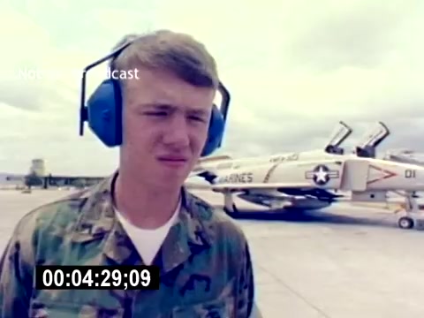 The still features a young serviceman in military camo attire wearing blue headphones, standing on an airfield. In the background, military aircraft are visible, likely part of USMC operations. The atmosphere suggests a focus on aviation ordnance and the recruiting theme of the film. The image captures a moment during a procedure or demonstration, emphasizing the individual's role in aviation operations.