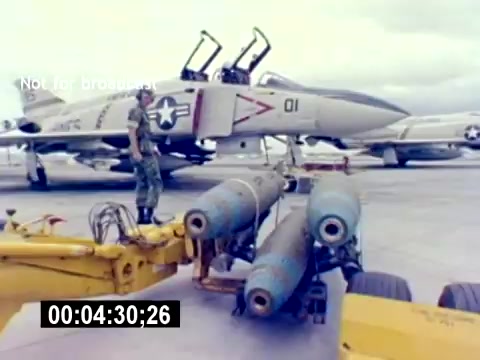 The 16mm still from the 'USMC Aviation Ordnance Recruiting Film - Silent' depicts a U.S. Marine Corps serviceman standing beside a loading vehicle equipped with two bombs. In the background, an F-4 Phantom fighter jet is visible, indicating an aviation setting. The scene underscores the themes of military operations and ordnance handling within a recruiting context.
