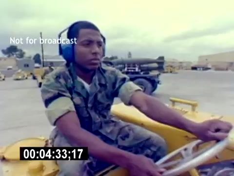 The still features a young man in military camouflage, wearing headphones, operating what appears to be a small, yellow vehicle in a military setting. The background includes various military equipment, suggesting a focus on aviation ordnance operations. The image captures a moment of concentration and duty, likely intended to highlight the responsibilities of USMC Aviation Ordnance personnel. The timestamp indicates the specific moment within the film.