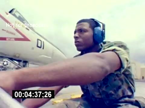 The still features a young Marine wearing a camouflage uniform and blue headphones, focused intently on operating equipment near a military aircraft. The aircraft, possibly a jet, is marked with the code 'D1.' The scene suggests an atmosphere of precision and dedication, typical of military training environments. The overall context relates to aviation ordnance and emphasizes the responsibilities of Marines in this specialized field.