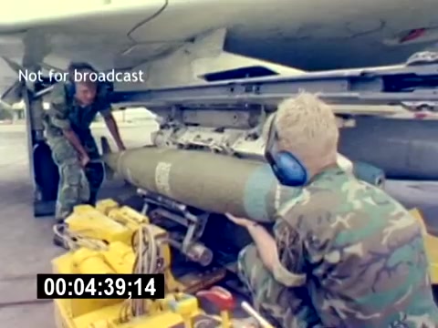 The still depicts a scene from the USMC Aviation Ordnance Recruiting Film, focusing on two individuals in military attire. One serviceman is engaged in the process of loading a bomb onto an aircraft, while another assists nearby. The environment appears to be an airfield, with equipment visible in the foreground. The image conveys a sense of teamwork and technical skill within the context of military aviation operations.