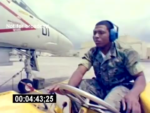 The still depicts a service member operating a small ground vehicle on an airfield, likely involved in aviation ordnance operations. The individual wears camouflage clothing and headphones, suggesting they are in a busy environment near military aircraft. In the background, an aircraft is visible, indicating the film's focus on the USMC Aviation Ordnance field. The scene captures a moment of activity and professionalism in military aviation operations.