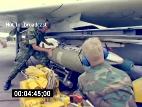 The still features two U.S. Marine Corps personnel in camouflage uniforms, actively loading a large ordnance onto an aircraft. One individual is positioned near the front, guiding the weapon, while the other is closer to the base, using mechanical equipment for assistance. Brightly colored machinery is visible in the foreground. The scene captures the technical and operational aspects of aviation ordnance, highlighting teamwork and precision in military operations. The 'Not for broadcast' label indicates that this material is for internal use only.