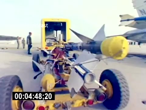 The still shows a close-up view of an aircraft towing vehicle designed to transport munitions. It features a large missile mounted on a wheeled chassis, with several mechanical components visible. In the background, military personnel, likely USMC members, are gathered near an aircraft. The setting appears to be a military airfield, demonstrating aspects of aviation ordnance operations.