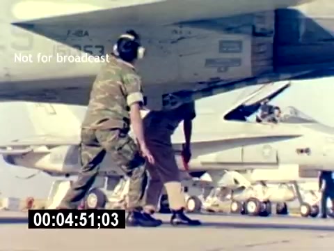 The still from the 'USMC Aviation Ordnance Recruiting Film - Silent' shows two military personnel working underneath an aircraft. One individual in a military uniform is positioned to the left, while another, dressed in beige, is visible beneath the aircraft's wing. The aircraft itself is likely a jet fighter, and there are additional jets parked in the background. The image captures a moment of teamwork and technical activity in an aviation context.