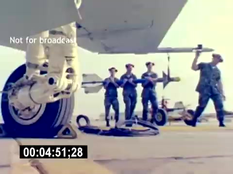 The still from the 'USMC Aviation Ordnance Recruiting Film - Silent' shows a military scene on an airfield. Prominently featured is a close-up of an aircraft wheel, while in the background, a group of service members is handling a missile or ordnance. They are dressed in military uniforms, indicating their role in aviation ordnance. The setting reflects the operational environment of the U.S. Marine Corps, showcasing teamwork and technical proficiency.