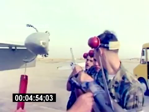 The still from the 'USMC Aviation Ordnance Recruiting Film - Silent' shows a group of U.S. Marine Corps personnel engaged in a training or operational activity related to aviation ordnance. Two individuals are prominently featured, wearing protective headgear and focusing on loading or handling ammunition or equipment. The background suggests a military airfield setting with aircraft visible, highlighting the specialized role of aviation ordnance in the Marine Corps. The image captures a moment of teamwork and technical operation without any accompanying audio.