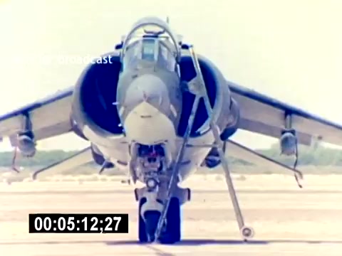 The still features a military aircraft, likely a Harrier jet, parked on an airfield. The focus is on the front of the aircraft, showcasing its fuselage and cockpit design. The environment appears to be a sunny day, with a clear sky and a barren landscape in the background. This frame reflects the aviation and technical aspects of the USMC Aviation Ordnance program, emphasizing the aircraft's capabilities.