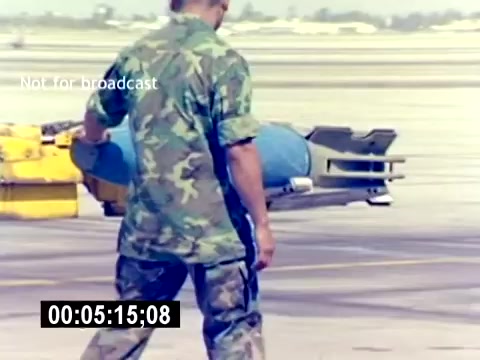 The still features a United States Marine Corps (USMC) service member in a camouflage uniform, handling a blue ordnance item—likely a bomb or missile—on an airstrip. In the background, yellow equipment and a clear sky can be seen, indicating a busy military aviation environment. The image captures a moment focused on the operational aspect of aviation ordnance handling, emphasizing the responsibilities of Marines in this field.