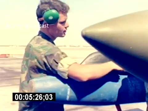 The still depicts a young Marine wearing camouflage fatigues and ear protection, focused on handling a missile on a military aircraft. The background shows an airfield, suggesting an operational setting for aviation ordnance work. The image captures a moment emphasizing the Marine's role in maintaining aviation readiness.