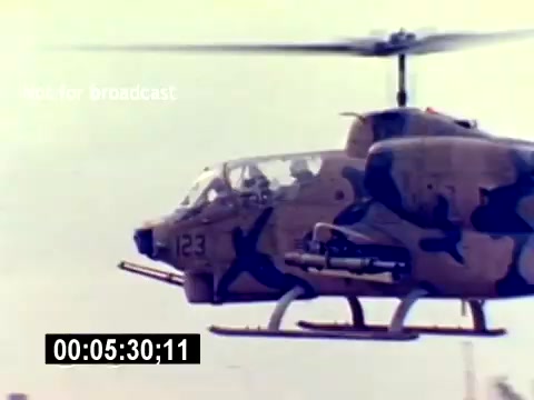 The still features a military helicopter, specifically one that appears to be the AH-1 Cobra, showcasing its striking camouflage paint. The helicopter is in flight, captured from a lateral angle. You can see the cockpit with visible instrumentation and the distinctive shape of the rotor blades creating motion. The film is part of a USMC Aviation Ordnance Recruiting effort, emphasizing the role of aviation and ordnance within the Marine Corps. No soundtrack accompanies this image, as it is a silent film.
