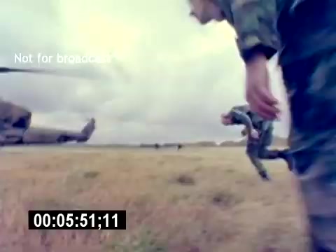 The still from the 'USMC Aviation Ordnance Recruiting Film - Silent' shows military personnel engaged in activities near a helicopter. The focus is on their actions as they move across a grassy area, suggesting a training or operational scenario. The environment appears open, with clouds in the sky, indicating possibly overcast weather. A timestamp at the bottom suggests the sequence is part of a larger film, emphasizing the action and movement inherent in military operations.