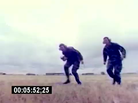 The still shows two individuals in military attire, likely USMC personnel, running across a field. They appear focused and determined, with a cloudy sky in the background. The image captures a moment emphasizing physical activity and teamwork, typical of a recruiting film for aviation ordnance. The timestamp at the bottom indicates the exact moment in the film.