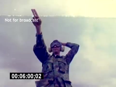 The still features a Marine in camouflage uniform, wearing protective ear muffs and goggles. He is standing outdoors, possibly on an airfield, with his arms raised in a signaling gesture. The sky appears cloudy or smoky, adding a dramatic backdrop to the scene. The frame includes a timestamp and a 'Not for broadcast' overlay, indicating it is part of a film intended for training or recruiting purposes rather than public viewing.