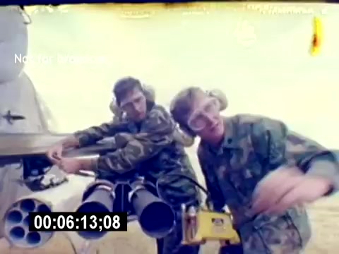 The still from the 'USMC Aviation Ordnance Recruiting Film - Silent' shows two servicemen in military camouflage uniforms working on an aircraft armament system. They appear focused, with one operating a piece of equipment while the other assists, all in a military environment. The background suggests a grassy or desert area, indicating an outdoor setting for the ordnance operations. The lack of a soundtrack highlights the visual emphasis on their activities and teamwork.