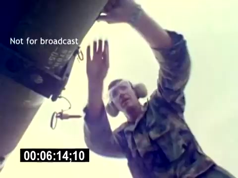 The still captures a U.S. Marine Corps aviation ordnance worker, shown from a low angle, interacting with military equipment. The individual is wearing protective earmuffs and a camouflaged uniform, indicating a focus on safety and professionalism. The scene emphasizes the hands-on nature of ordnance handling, showcasing the worker's engagement with the task at hand. The muted colors and military context reinforce the organized and serious atmosphere typical of training or recruitment materials.