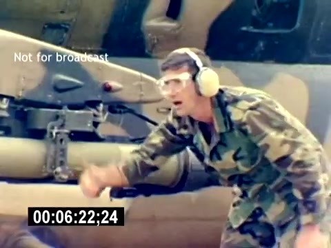 The still depicts a uniformed Marine, wearing protective headphones, gesturing emphatically in front of a military helicopter. His camouflaged outfit reflects a typical field environment. The background features the helicopter's underside, highlighting its mechanical elements, suggesting a focus on aviation ordnance operations. The lack of sound emphasizes the visual communication of commands or instructions in a military setting. The timestamp indicates it's around 6 minutes into the film.