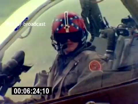 The still from the 'USMC Aviation Ordnance Recruiting Film - Silent' features a pilot inside an aircraft cockpit. The pilot is wearing a helmet adorned with a distinctive red and blue design, indicative of military aviation. The cockpit is equipped with various instruments and controls, suggesting an operational setting. The background appears to show a blurred aerial landscape, emphasizing the aircraft's altitude and movement. The overall tone conveys a sense of professionalism and readiness associated with USMC aviation.