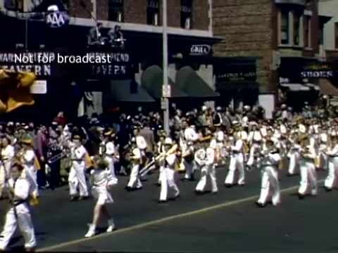 The still captures a scene from the Holland, Michigan Tulip Festival Parade in the late 1940s. It features a marching band in uniform, playing instruments as they parade down a street lined with onlookers. Spectators watch from the sidewalks, while buildings and storefronts are visible in the background, reflecting the community atmosphere of the event. The vibrant colors of the band uniforms and the festive ambiance convey the celebratory spirit of the festival.