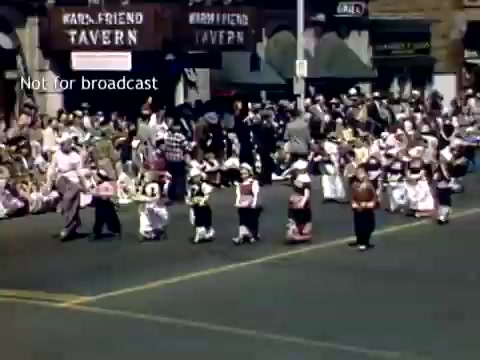The still from the 'Holland, Michigan Tulip Festival Parade' in the late 1940s showcases a vibrant scene with a group of children dressed in traditional Dutch attire, participating in the parade. In the background, a large crowd of spectators gathers along the street, and buildings, including a tavern, can be seen. The atmosphere suggests a festive celebration, highlighting the cultural significance of the tulip festival.