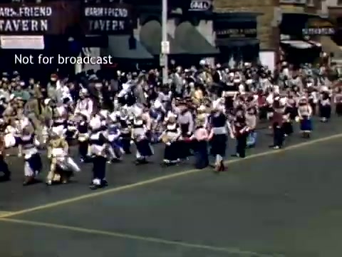 The still captures a vibrant scene from the Holland, Michigan Tulip Festival Parade in the late 1940s. A lively procession moves down the street, with participants dressed in traditional attire, showcasing cultural heritage. Spectators line the sidewalks, while storefronts in the background hint at a bustling local atmosphere. The image conveys a festive and communal spirit typical of parades during that era.
