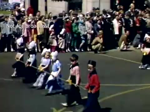 The still captures a vibrant scene from the Holland, Michigan Tulip Festival Parade in the late 1940s. In the foreground, several children dressed in traditional attire are marching, some holding ropes or sticks. They appear to be participating in a festive procession. A large crowd of onlookers can be seen in the background, dressed in various colorful outfits, reflecting the lively atmosphere of the event. The scene conveys a sense of community celebration around the cultural significance of tulips.