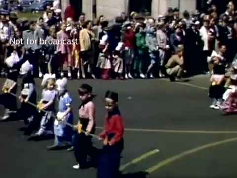 The still captures a lively scene from the Holland, Michigan Tulip Festival Parade in the late 1940s. Children, dressed in colorful attire, participate in the parade, holding props like sticks or flags. A large crowd of onlookers, dressed in a mix of historical fashions, lines the street, enjoying the festive atmosphere. The background features a mix of trees and buildings, adding to the charm of the event.