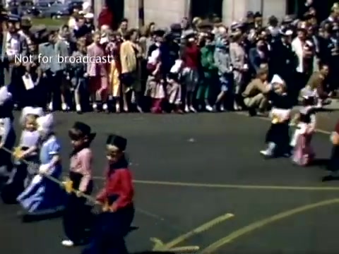 The still captures a vibrant scene from the late 1940s Holland, Michigan Tulip Festival Parade. Children dressed in traditional attire march along the street, holding sticks, while a lively crowd observes from the sidelines. The spectators, dressed in a mix of colorful clothing, suggest a festive atmosphere celebrating the parade and the festival's Dutch heritage.