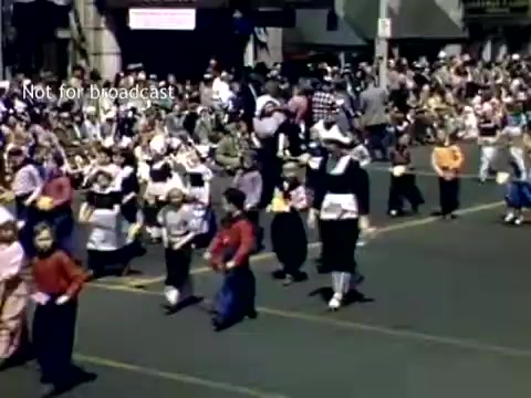 The still shows a vibrant scene from the Tulip Festival Parade in Holland, Michigan, during the late 1940s. Groups of children in traditional costumes march along a street lined with onlookers. The festive atmosphere is enhanced by colorful attire, drawing attention to the celebration of Dutch heritage and culture. The crowd in the background adds to the lively ambiance of the event.