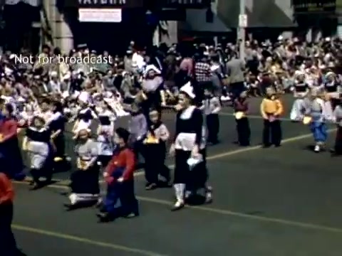 The still from the 'Holland, Michigan Tulip Festival Parade' in the late 1940s captures a vibrant scene of participants dressed in traditional Dutch costumes. A group of children can be seen parading, with some in black pants and colorful tops, while others wear bonnets and dresses. The parade is bustling with a large crowd of onlookers in the background, reflecting a festive atmosphere. The event celebrates Dutch heritage, emphasizing community and tradition.