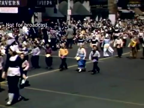 The still captures a lively scene from the late 1940s Holland, Michigan Tulip Festival Parade. Rows of participants, including children in colorful traditional costumes, march along a street lined with spectators. The backdrop features businesses, creating a festive and community-oriented atmosphere, highlighting the celebration's cultural significance.