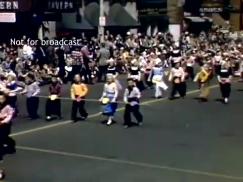 The still captures a vibrant scene from the late 1940s Holland, Michigan Tulip Festival Parade. In the foreground, a diverse group of parade participants walks in colorful attire, showcasing traditional dress and joyful expressions. The background is filled with a large crowd of spectators, many dressed in casual 1940s fashion, along with storefronts visible behind them. The overall atmosphere is festive, reflecting the celebration of Dutch heritage through music and dance.