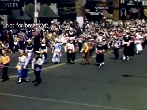 The still captures a vibrant scene from the late 1940s Holland, Michigan Tulip Festival Parade. A colorful procession features groups of dancers in traditional attire, showcasing cultural heritage. Crowds line the streets, eagerly watching the festivities, while storefronts and signs add to the lively atmosphere. The image conveys a sense of community celebration and joy.