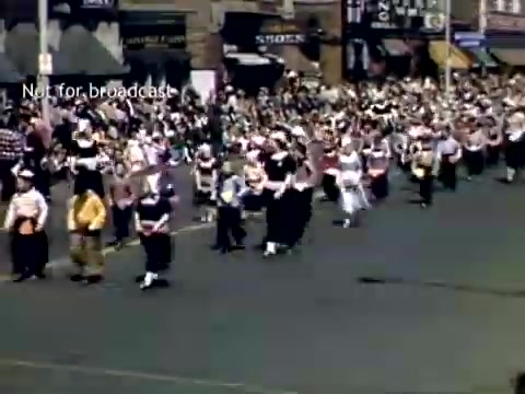 The still captures a vibrant scene from the late 1940s Holland, Michigan Tulip Festival Parade. A large group of participants dressed in traditional attire marches along a street lined with spectators. The atmosphere is festive, showcasing cultural heritage with colorful costumes and community involvement. Well-known storefronts and a lively crowd add to the parade's celebratory context.