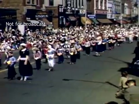 The still captures a lively scene from the Holland, Michigan Tulip Festival Parade in the late 1940s. A large group of participants, dressed in traditional Dutch attire, marches along a street lined with spectators. The crowd, consisting of men, women, and children, watches the parade, which showcases the festive atmosphere of the event. Shops and storefronts are visible in the background, contributing to the historic vibe of the scene.