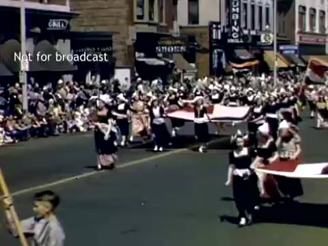 The still captures a lively scene from the Holland, Michigan Tulip Festival Parade in the late 1940s. It features a group of marchers dressed in traditional Dutch costumes, some carrying a large flag. A crowd of spectators lines the street, with children and adults enjoying the festive atmosphere. Shops and storefronts can be seen in the background, framed by a clear sky.