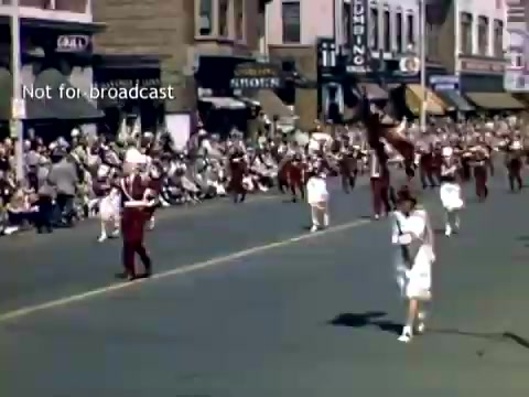 The still depicts a lively scene from the Holland, Michigan Tulip Festival Parade in the late 1940s. Participants dressed in colorful uniforms march down a bustling street, flanked by onlookers gathered along the sidewalks. Buildings and storefronts from the era are visible in the background, creating a festive atmosphere enriched by the historic context of the parade.