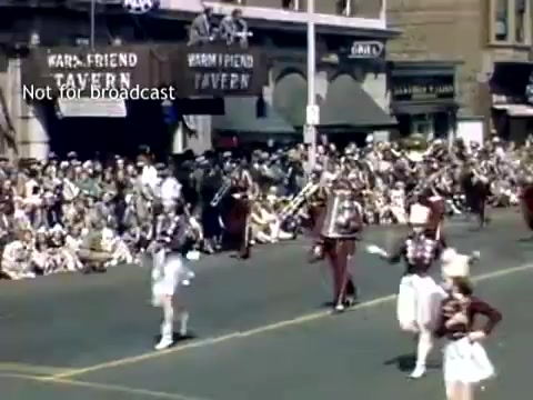 The still captures a vibrant scene from the late 1940s Holland, Michigan Tulip Festival Parade. A marching band dressed in uniforms is visible, accompanied by performers, likely dancers, in elaborate costumes. The crowd gathers along the street, with buildings in the background advertising 'Warm Friend Tavern.' The festive atmosphere reflects the celebration of the annual tulip festival, showcasing community spirit and tradition.