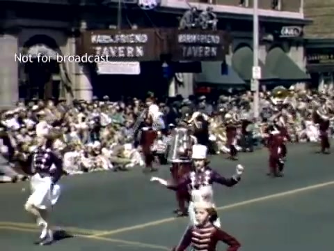 The still captures a vibrant scene from the late 1940s Holland, Michigan Tulip Festival Parade. In the foreground, a marching band in uniforms is performing, accompanied by young performers, while a large crowd watches from the sidelines. The setting features historic buildings, including a tavern, contributing to the festive atmosphere of the event. Bright colors and enthusiastic expressions convey the celebration of the festival.