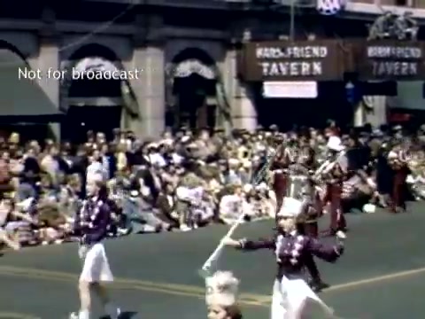 The still depicts a lively scene from the Holland, Michigan Tulip Festival Parade in the late 1940s. Marching performers in colorful uniforms, likely a marching band, are leading the procession. Spectators line the street, seated and enjoying the festivities, while buildings adorned with banners, including the 'HARD-FRIEND TAVERN,' provide a backdrop to the event. The atmosphere is festive and community-oriented, showcasing local traditions and parades.