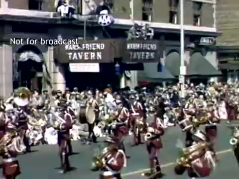 The still captures a vibrant scene from the late 1940s Holland, Michigan Tulip Festival Parade. In the foreground, a marching band in red uniforms plays instruments while parading down a street lined with spectators. The background features a tavern with large signage and a lively crowd gathered on the sidewalks, all contributing to the festive atmosphere typical of the event.